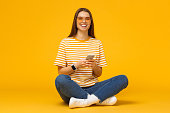 Beautiful smiling woman sitting on floor with legs crossed and holding mobile phone, isolated on yellow background