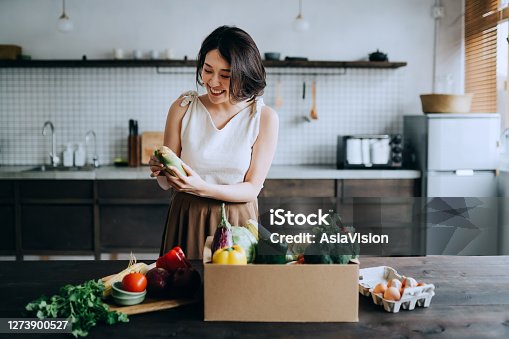istock Beautiful smiling young Asian woman received a full box of colourful and fresh organic groceries ordered online by home doorstep delivery service. She is sorting out the groceries and preparing to cook a healthy meal 1273900527