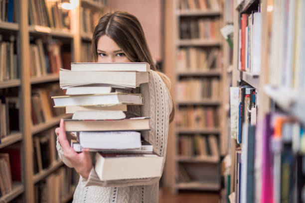 Beautiful teenage girl hiding behind a stack of books Beautiful teenage girl hiding behind a stack of books literature stock pictures, royalty-free photos & images