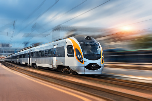 Beautiful train in motion at the railway station at sunset in Europe. Modern intercity train on the railway platform with motion blur effect. Industrial landscape with passenger train on railroad