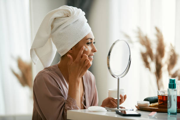 Beautiful woman applying face cream while looking herself in a mirror at home. Young happy woman looking herself in a mirror and using moisturizer on her face skin at home. applying face cream stock pictures, royalty-free photos & images