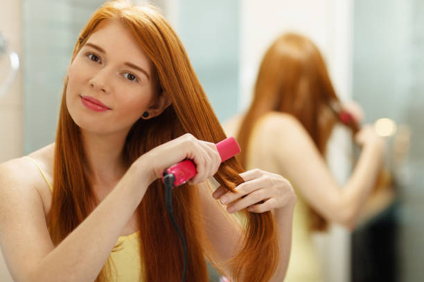 Beautiful redhead girl using electric hair straightener in bathroom....