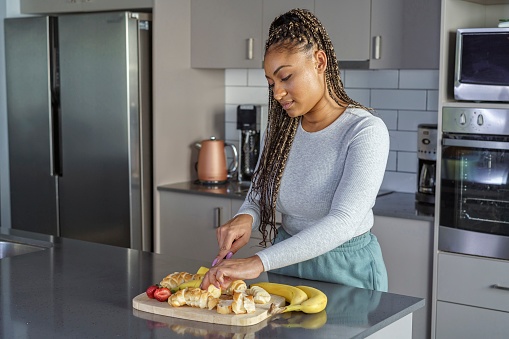 A beautiful black woman stands in her kitchen and prepares a delicious meal. She is slicing bread and chopping fresh fruit. Simple pleasures, routine, healthy eating and domestic life concepts.