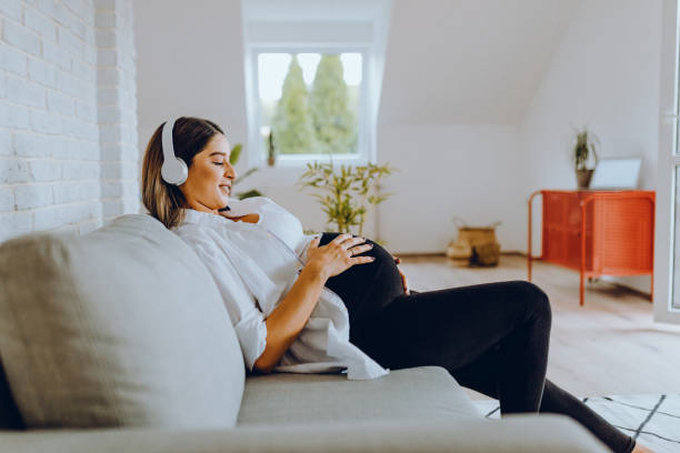 beautiful woman sitting on the couch at home enjoying pregnancy with laptop and music stock photo