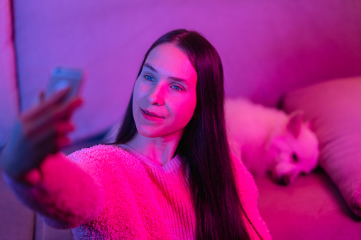 A beautiful woman is taking selfies with a smart phone with her dog in pink and blue lit room.