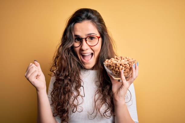 Beautiful woman with curly hair holding bowl with healthy peanuts over yellow background screaming proud and celebrating victory and success very excited, cheering emotion Beautiful woman with curly hair holding bowl with healthy peanuts over yellow background screaming proud and celebrating victory and success very excited, cheering emotion nut food stock pictures, royalty-free photos & images