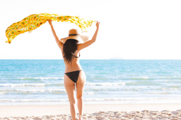 Beautiful women wearing black bikini standing and sunhat with her hand holding a yellow scarf on tropical beach in summer day, sea beach of tropical island. stock photo