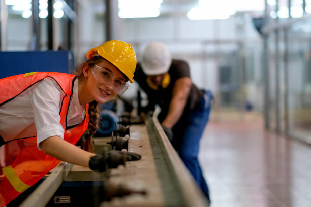 Beautiful worker or technician or engineer woman smile and look forward in front of rail of the machine with her co-worker as background in factory Beautiful worker or technician or engineer woman smile and look forward in front of rail of the machine with her co-worker as background in factory. trainee stock pictures, royalty-free photos & images
