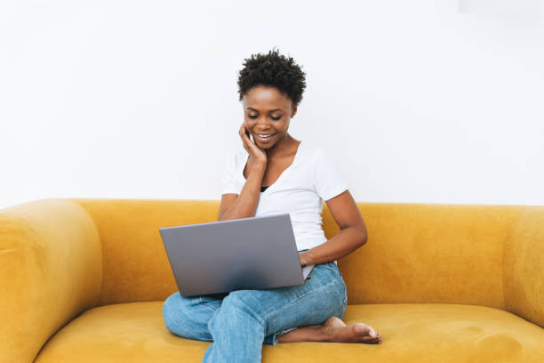 Beautiful young African American woman in white t-shirt and blue jeans using laptop sitting on yellow sofa in the bright modern interior stock photo