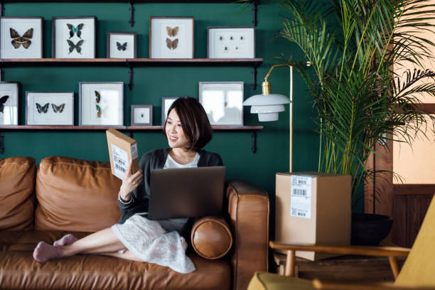 beautiful young asian woman relaxing on sofa at home, shopping online with laptop. she is holding a delivered parcel, can't wait to unbox the purchases. online shopping, enjoyable customer shopping experience - unbox stockfoto's en -beelden