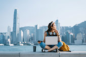 istock Beautiful young Asian woman sitting cross-legged by the promenade, against urban city skyline. She is wearing headphones around neck, using smartphone and working on laptop, with a coffee cup by her side. Looking away in thought. Lifestyle and technology 1319723972