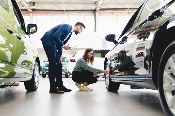 Beautiful young caucasian female client customer choosing new car, trying checking its options, tire, wheels while male shop assistant helping her to choose it at dealer auto shop stock photo