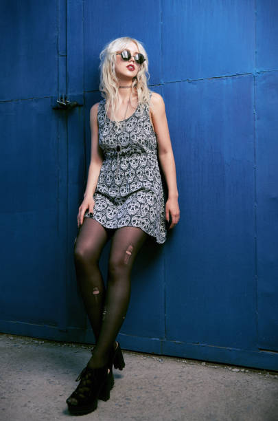 Beautiful young grunge (rock) girl in dress, ripped pantyhose and sunglasses standing at wall. Full length outdoor portrait of informal model stock photo