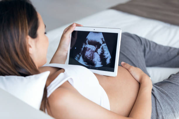 Beautiful young pregnant woman looking ultrasound of her baby on digital tablet while lying on bed. Shot of beautiful young pregnant woman looking ultrasound of her baby on digital tablet while lying on bed. obstetrician photos stock pictures, royalty-free photos & images