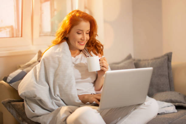 Beautiful young red haired smiling woman working on laptop and drinking coffee while sitting in a big comfortable couch covered with blanket over shoulders at home stock photo