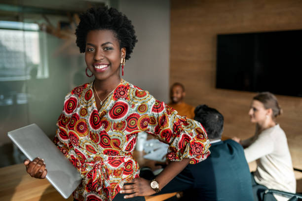 bella giovane donna d'affari africana nera sorridente che tiene il laptop, i colleghi tengono una riunione in background - business woman foto e immagini stock