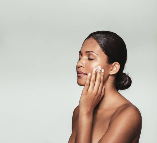 Beautiful young woman applying moisturizer to her face Beautiful young woman with eyes closed while applying moisturizer to her clean face. Close up portrait female model applying face cream against grey background. applying face cream stock pictures, royalty-free photos & images