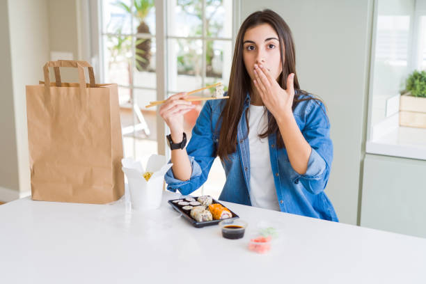 Beautiful young woman eating asian sushi from home delivery cover mouth with hand shocked with shame for mistake, expression of fear, scared in silence, secret concept Beautiful young woman eating asian sushi from home delivery cover mouth with hand shocked with shame for mistake, expression of fear, scared in silence, secret concept how do you say shut up in japanese stock pictures, royalty-free photos & images