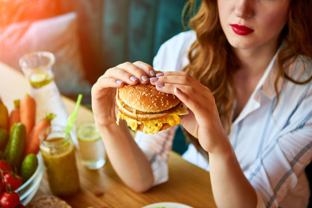 Beautiful young woman eating hamburger instead of salad in kitchen. Cheap junk food vs healthy diet stock photo