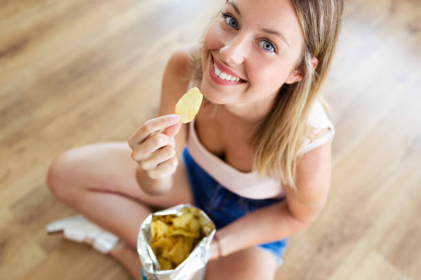 schöne junge frau, die kartoffeln zu essen, während auf dem boden zu hause sitzen. - chips potato stock-fotos und bilder