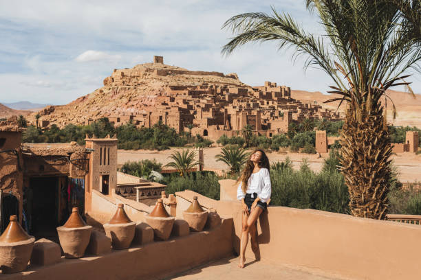 Beautiful young woman happy to travel in Morocco. Ait-Ben-Haddou kasbah on background. Wearing in white shirt and jeans shorts. stock photo