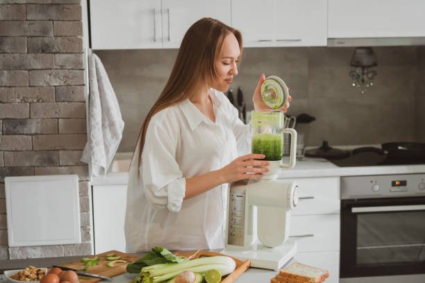 beautiful young woman preparing healthy green smoothie in her kitchen stock photo