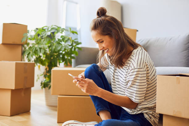 Beautiful young woman texting while moving to new apartment Beautiful young woman texting while moving to new apartment relocation stock pictures, royalty-free photos & images