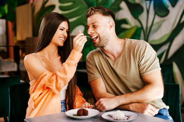Beautiful young woman with a smile on her face feeds her boyfriend with chocolate cake in a cafe stock photo