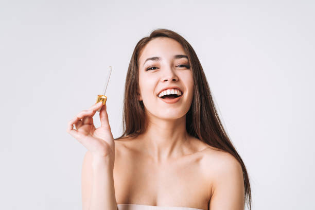 Beauty portrait of happy smiling asian woman with dark long hair with pipette with organic oil on clean fresh skin face and hands on white background isolated stock photo