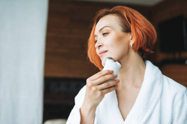 Beauty portrait of smiling woman 35 year in white bathrobe with clean fresh face and hands with red hair doing fasial massage with microcurrent facial massager at bath room, treat yourself, home body care stock photo