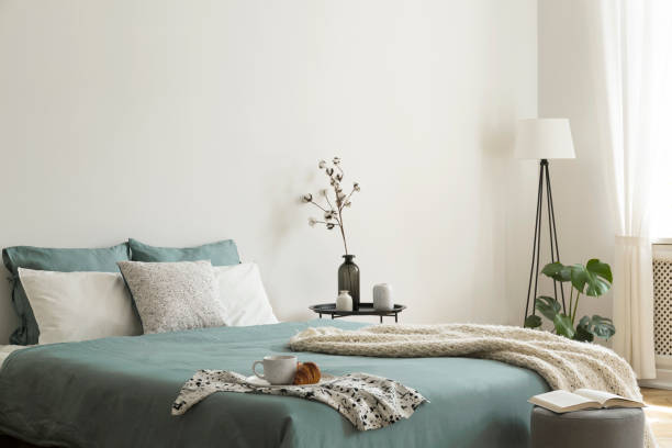 Bedroom interior with sage green and white sheets and cushions and a blanket. Black metal table with vases beside the bed. A lamp standing in the corner. Real photo. stock photo