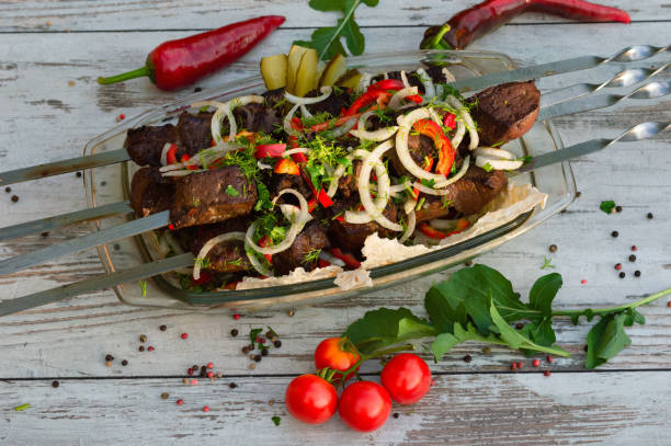 Beef liver on trochantel is served in a glass large plate on Lavashe with peppers and cherry tomatoes. On a wooden grated background. Picnic. Bright background with liver, close up Beef liver on swords, pre marinated in wine and spices, fried on open fire. Family holiday in nature. Proper nutrition. Beef Liver Grilled Recipe stock pictures, royalty-free photos & images