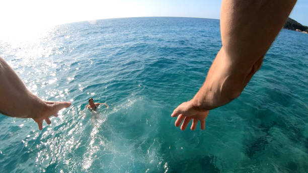 POV behind man's arms as he jumps off cliffs over sea Young woman is below swimming cliff jumping stock pictures, royalty-free photos & images