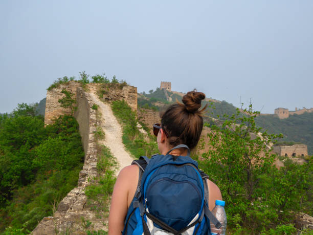 Beijing - A woman with hiking backpack walking around a unrenewed Gubeikou part of Great Wall of China. The wall is spreading A woman with hiking backpack walking around a unrenewed Gubeikou part of Great Wall of China. The wall is spreading on tops of mountains. Dense forest around it. World wonder. Tradition and history mutianyu stock pictures, royalty-free photos & images