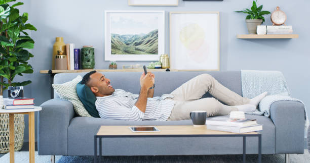 Being at home is the best feeling Full length shot of a handsome young man lying down on the sofa in his home and using his cellphone lying down stock pictures, royalty-free photos & images