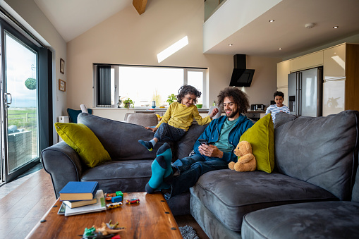 Two children playing near the sofa where their father is sitting while on a staycation at a lodge in the Northeast of England together. He is using his mobile phone while one of the boys jumps over the chair to sit with him.