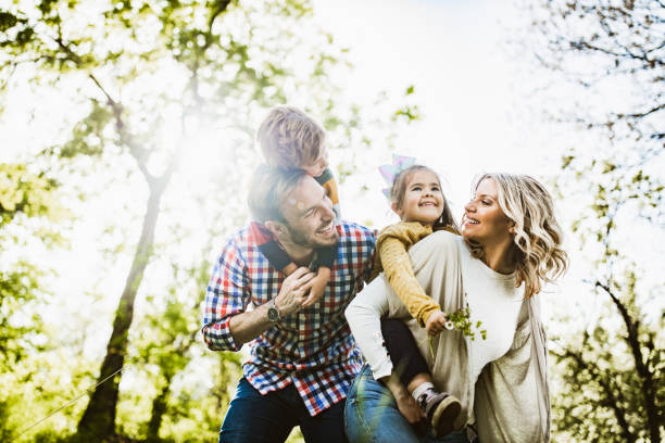Below view of playful family having fun while piggybacking in nature. Low angle view of happy parents having fun while piggybacking their small kids in nature. family outside stock pictures, royalty-free photos & images