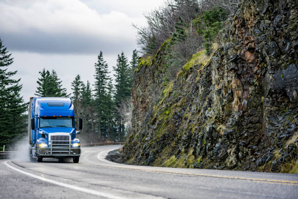 Big rig blue semi truck with grill guard transporting cargo in semi trailer running on the winding wet road with rain dust with rock cliff on the side stock photo