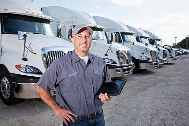 Big rig trucks behind man holding tablet A truck driver is standing in a large parking lot in front of several large trucks.  He is facing the camera, and he is wearing a short-sleeved blue shirt, blue jeans, a brown belt and a gray hat.  He is holding a black tablet with his left hand.  There are six big rigs seen lined up behind him, going from left to right.  The large trucks all look the same, being all white and having silver grills and mirrors.  There are multiple cars seen parked to the right of the trucks.  A cloudy blue sky is seen in the background. truck driver stock pictures, royalty-free photos & images