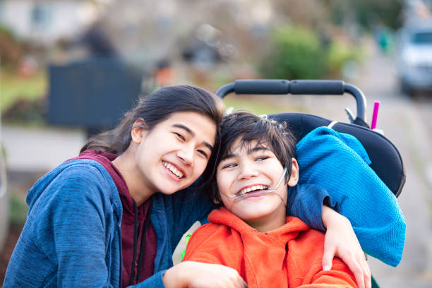 Big sister hugging disabled brother in wheelchair outdoors, smiling stock photo