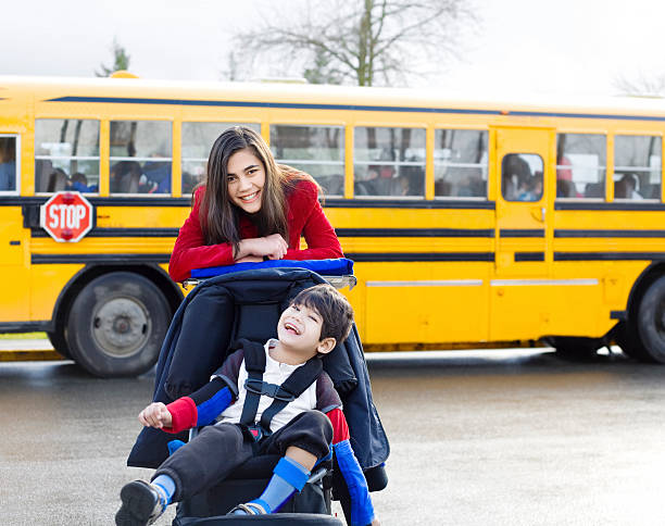 Big sister with disabled brother in wheelchair by school bus stock photo