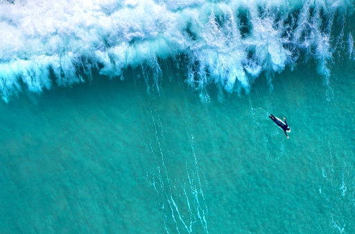 Big sea wave splashing with foam while a lonely surfer wearing a diving suit is seen from above swimming to calm waters. Ocean view from above, drone point of view.