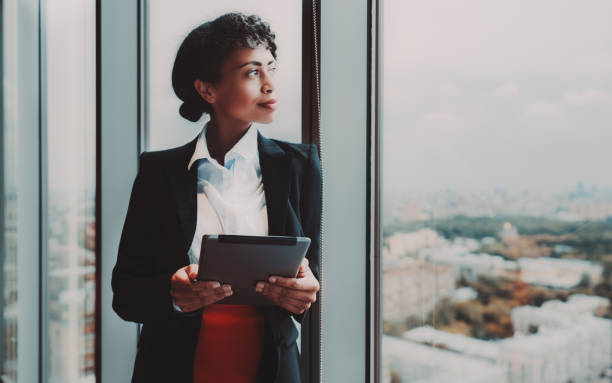 Biracial businesswoman and tablet pc A dazzling young African-American woman entrepreneur with a digital tablet in her hands is standing near a huge panoramic window indoors of a business high-rise and pensively looking at the cityscape ceo stock pictures, royalty-free photos & images