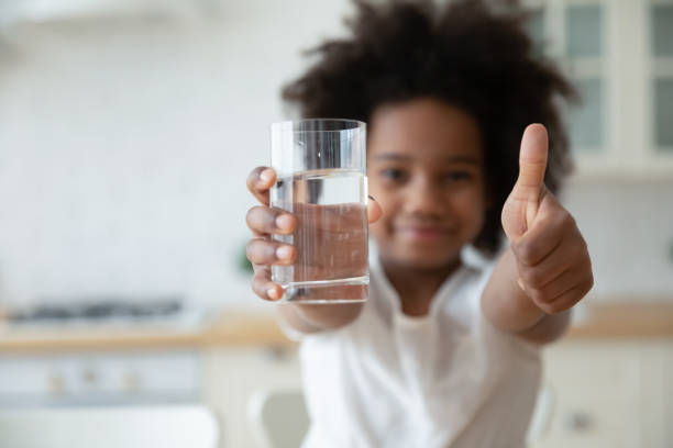 Biracial girl holding glass of water, showing thumbs up gesture. Focus on happy small african american girls hands holding glass with fresh pure water and showing thumbs up gesture. Smiling little biracial kid recommending healthcare habit, morning refreshment. drinking water stock pictures, royalty-free photos & images