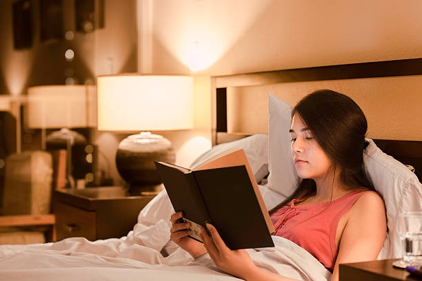 Biracial teen girl reading book  in bed at night stock photo