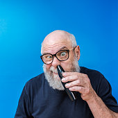 A funny, nerdy, vain senior adult man with a bizarre, exaggerated facial expression is staring at the camera through his geeky black rimmed glasses as he carefully pokes a portable electric razor with a specialized circular nose hair clipping attachment up deeply into his nostril to trim his nose hair. He's sitting in front of a dark blue background wall.