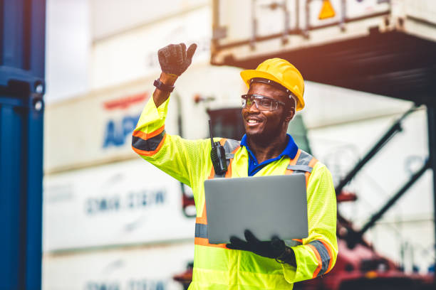 Black African happy worker working in logistic shipping with laptop computer control loading containers at port cargo. Import export shipping industry people. stock photo