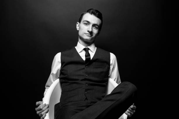 Black and white portrait of a young handsome man in a white shirt, black tie and vest, sitting and looking at the camera Black and white portrait of a young handsome man in a white shirt, black tie and vest, sitting and looking at the camera, against plain studio background arrogance stock pictures, royalty-free photos & images