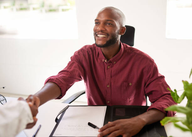A Black businessman greets a client from his office desk for in person meeting stock photo