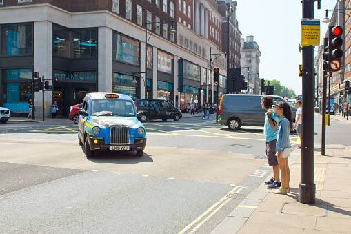 Black cab with advertising on Regent Street, London with shoppers/tourists waiting to cross at a junction.